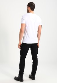 Witte katoenen t-shirt gecombineerd met zwarte cargobroek, voorzien van zijzakken en een slim fit, gezien van achteren. Zwarte laarzen maken de look compleet.