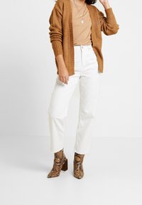 Brun stickad kofta, beige topp, vita jeans med raka ben och bruna ormskinnsankelstövlar med blockklackar. Mot en enfärgad bakgrund.