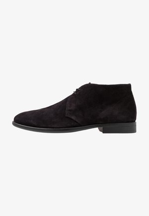 Bottines en daim noir avec une texture lisse, un bout rond et un laçage minimal. Elles présentent une semelle plate et des accents de couture subtils.