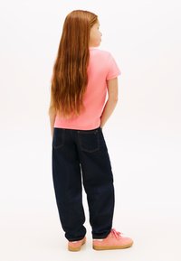 Παιδί με μακριά κόκκινα μαλλιά που φοράει ροζ μπλούζα, σκούρο άνετο τζιν και ροζ αθλητικά παπούτσια, στέκεται με τα χέρια στις τσέπες και κοιτάει πλάγια.