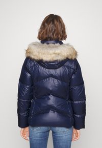 Mörkblå puffjacka med fuskpäls på huvan, med en kviltad design och en glänsande yta. Synliga sömmar och sidofickor.