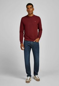 Bordeaux sweatshirt met ronde hals, slim fit; blauwe denim jeans; witte sneakers met bruine accenten; overal een gladde textuur.