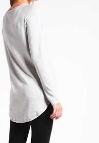 Tunika a maniche lunghe grigia realizzata in tessuto morbido, con orlo curvo, caratterizzata da una vestibilità comoda e una texture liscia. Vengono indossati leggings neri.