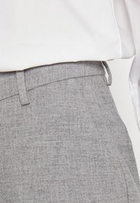 Grijze textuurbroek met een gladde tailleband en riemlussen, gedeeltelijk bedekt door een witte blouse. Gedetailleerd met subtiele stofpatronen.