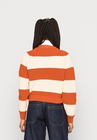 Sweter dzianinowy w szerokie pomarańczowe i kremowe paski, okrągły dekolt, ściągacze przy mankietach i u dołu. Nosi się go na ciemnoniebieskich spodniach dżinsowych. Pokazano widok z tyłu.