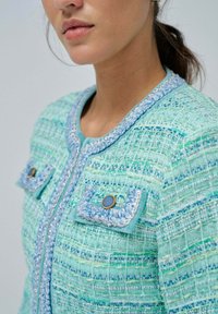 Giacca in tweed con texture in tonalità di blu e verde, caratterizzata da un collo rotondo, tasche decorate e rifiniture decorative lungo la zip.