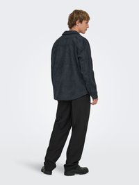 Chemise en daim noir avec un col à revers, manches longues et coupe décontractée, associée à un pantalon noir ample et des chaussures noires.