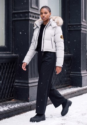 Jeune femme aux cheveux tressés marche sur un trottoir enneigé, portant une veste claire avec capuche en fourrure, un pantalon noir et des bottes noires près d’un bâtiment sombre.