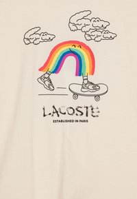 Beige tričko s barevným grafickým motivem skateboardisty, jehož nohy jsou tvořeny duhou a nad ním jsou mraky, s nápisem "LACOSTE" vytištěným níže.