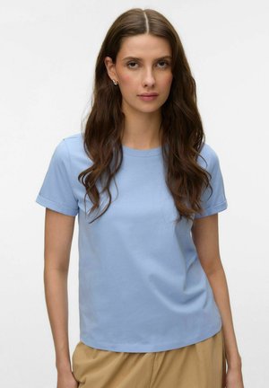 Woman with long brown hair wearing silver hoop earrings, a light blue short-sleeve t-shirt, and beige pants, facing forward against white background.