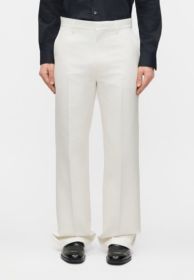 Άνδρας που φοράει εκρού παντελόνι με πιέτες, μαύρα επίσημα παπούτσια και μαύρο πουκάμισο με μακριά μανίκια, στέκεται μπροστά σε απλό φόντο.