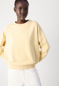 Gul överdimensionerad sweatshirt med rund halsringning och ribbade ärmavslut, kombinerad med vita jeans, med en diskret broderad logotyp på bröstet.