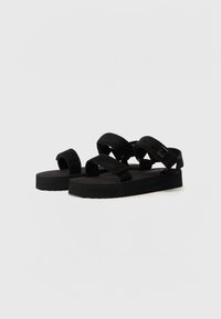 Sandales noires avec une semelle en caoutchouc épaisse et texturée ainsi que des bandes douces en suédé. Boucles réglables pour un maintien sécurisé. Design simple et minimaliste.