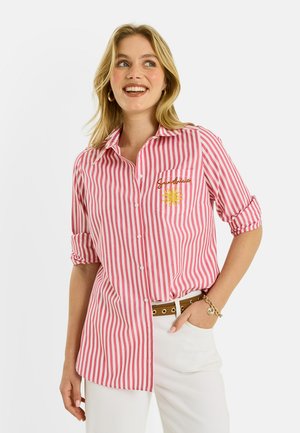 Mujer sonriente con camisa de rayas verticales rojas y blancas con botones, con texto "Sunshine" y bordado de sol, pantalones blancos y accesorios dorados.