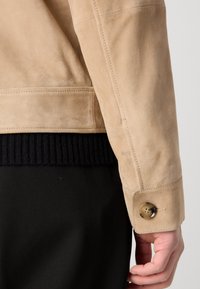 Beige suede jacket featuring a textured finish, ribbed black knit hem, and a brown button on the sleeve. Close-up of sleeve detail.