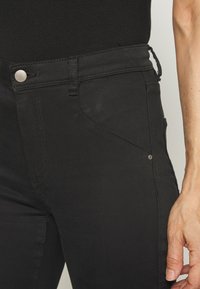 Jeans negros de denim con una textura suave. Presenta una costura decorativa en el bolsillo frontal y un botón plateado en la cintura.