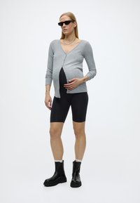 Femeie însărcinată purtând un cardigan gri, pantaloni scurți negri de ciclism, cizme negre și ochelari de soare, stând în picioare pe un fundal alb simplu.