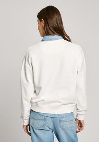 Sudadera blanca con un corte relajado, puños acanalados y dobladillo; superpuesta a una camisa de mezclilla azul clara, visible en el cuello.