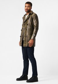 Olive green leather trench coat with double-breasted buttons, two side pockets, and a logo accent on the cuff, worn over denim jeans and black boots.