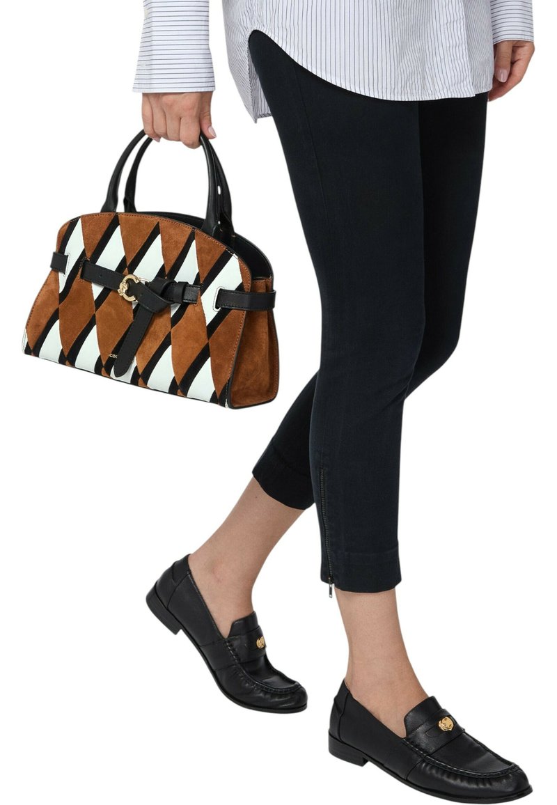 Borsa in suede marrone e bianca con motivi, dettagli neri, tenuta da una persona che indossa pantaloni neri corti e mocassini neri.