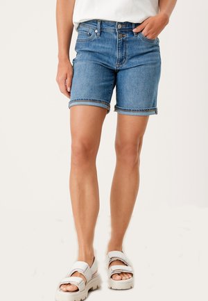 Femme portant un short en jean bleu retroussé, un top blanc et des sandales blanches, debout avec une main dans la poche sur un fond clair.