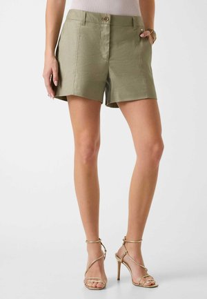 Mujer que lleva pantalones cortos de talle alto color verde oliva y sandalias doradas de tacón alto con tiras, de pie frente a un fondo claro y liso.