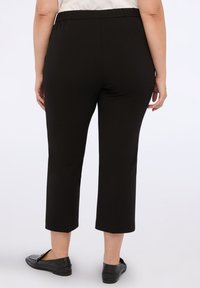 Kvinna som bär svarta cropped byxor och svarta loafers, stående med avslappnade händer vid sidorna mot en enkel vit bakgrund.