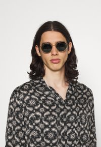 Lunettes de soleil rondes avec verres foncés, monture en métal doré. Chemise noire avec un motif paisley crème. Texture de tissu lisse.