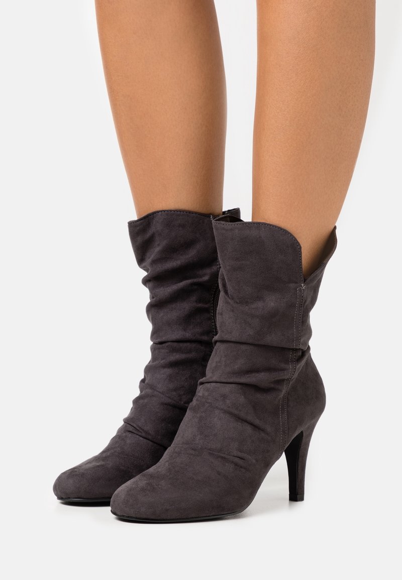 Bottes molles en suède gris foncé mi-mollet avec bouts pointus et talons hauts fins, portées sur des jambes nues sur un fond uni.