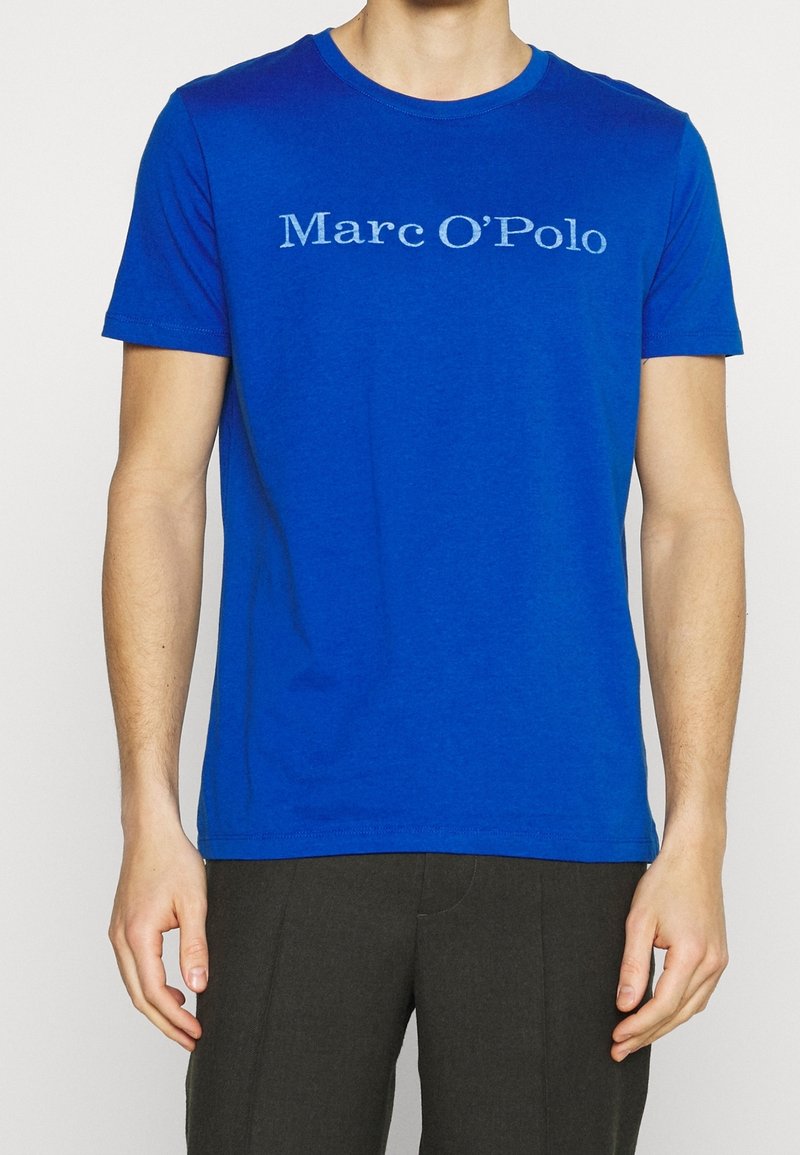 Homme portant un t-shirt à manches courtes bleu roi avec le texte blanc "Marc O’Polo" sur la poitrine et un pantalon gris foncé.