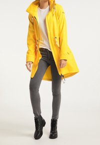 Kobieta w żółtym płaszczu przeciwdeszczowym, białej koszulce, szarych obcisłych dżinsach oraz czarnych sznurowanych botkach stojąca na białym tle.