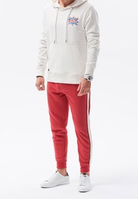 Felpa bianca con grafica "OMG", tasca a canguro; abbinata a pantaloni jogger rossi con strisce laterali e scarpe da ginnastica bianche.