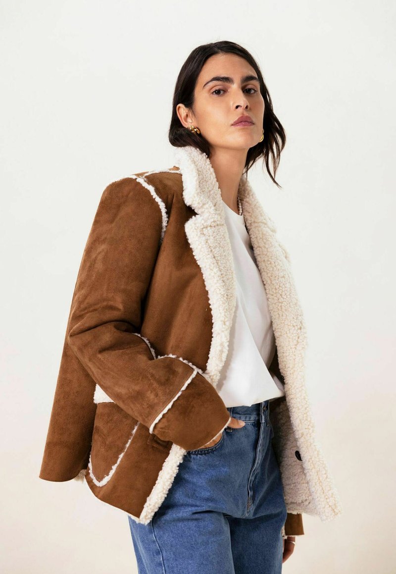 Veste en daim marron avec col et doublure en fausse fourrure blanche, présentant une couture contrastée, un design superposé, portée sur une chemise blanche et un jean bleu.