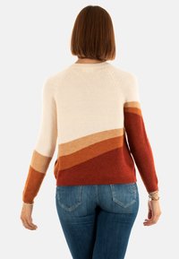Femme aux cheveux bruns mi-longs portant un pull coloré en crème, orange et rouge et un jean bleu, vue de dos.