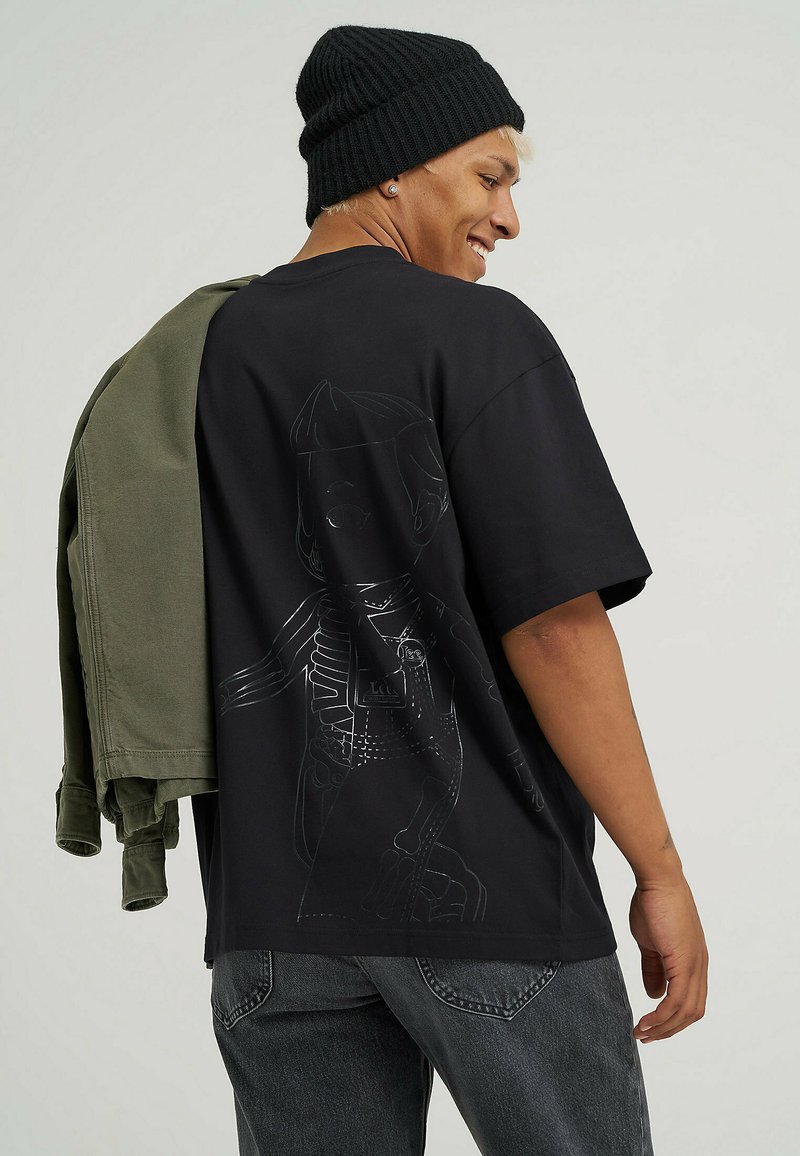 Sort bomuld t-shirt med en linjeteckning grafisk på ryggen. Casual pasform, parret med en olivengrøn jakke vist over den ene skulder.