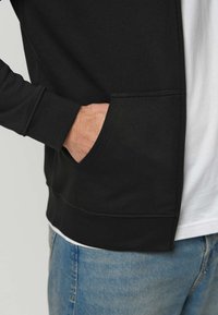 Melns sporta krekliņš ar priekšējo kabatu, teksturētu audumu un ribotām aprocēm, valkāts uz balta krekla un kombinēts ar gaiši ziliem džinsiem.