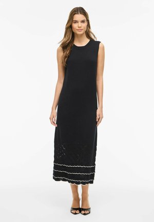 Kobieta stojąca w czarnej, bezrękawnikowej sukience maxi z wzorem diamentowym i falowanym dołem, w połączeniu z czarnymi czółenkami na odkryty palec na białym tle.