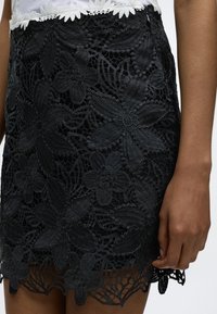 Μαύρη δαντελένια φούστα με μεγάλα λουλουδάτα σχέδια, με κυματιστό τελείωμα, συνδυασμένη με λευκό τοπ με λουλουδάτη δαντέλα στη μέση.