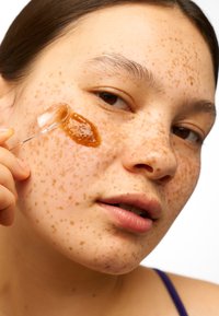 Translucent spatula applicerar en tjock, bärnstensfärgad ansiktsbehandling på fräknig hud. Bakgrunden är enkel vit, vilket framhäver produktapplikationen.