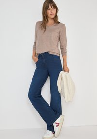 Blaue Jeans mit geradem Schnitt, kombiniert mit einem hellbraunen Langarmshirt. Weiße Sneaker mit roten Akzenten und einer cremefarbenen, gemütlichen Jacke.