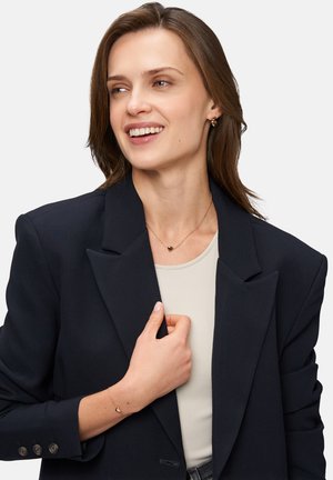 Zwarte blazer over een lichtgekleurde top, met een hartvormige ketting en een delicate armband. Soepele texturen en een elegante uitstraling.