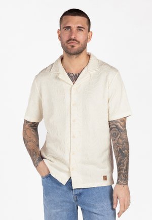 Mann mit kurzen Haaren und Ärmeltattoos, der ein strukturiertes cremefarbenes Kurzarmhemd und blaue Jeans trägt, steht mit einer Hand in der Tasche.