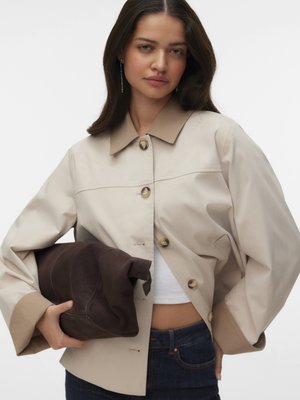 Femme aux cheveux foncés porte une veste beige par-dessus un haut blanc et un jean bleu, tenant un grand sac brun foncé plié sur fond blanc.