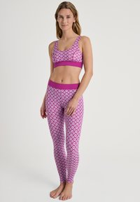 Vrouw staat blootsvoets in een bijpassende sport-bh en legging in roze en witte patronen tegen een effen lichte achtergrond.