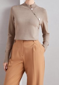 Beiger Pullover mit hohem Kragen und seitlichen Knopfverschlüssen, kombiniert mit hellbraunen, taillierten Hosen mit seitlichen Taschen und Falten.
