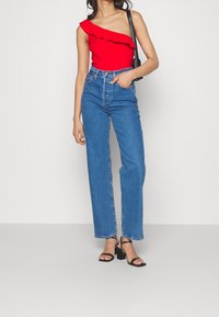Top rojo de un solo hombro con detalle de volantes, vaqueros azul de tiro alto y pernera recta, y sandalias negras de tacón cuadrado con tiras finas.