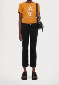 Mustard yellow t-shirt with white logo, paired with black cropped jeans. Black chunky sandals with buckle accents. Handheld black bag with chain.