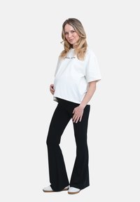 Vit bomullströja med texten "COOL MAMA", kombinerad med svarta utsvängda byxor. Modellen står med ena handen på magen och har på sig vita sneakers.