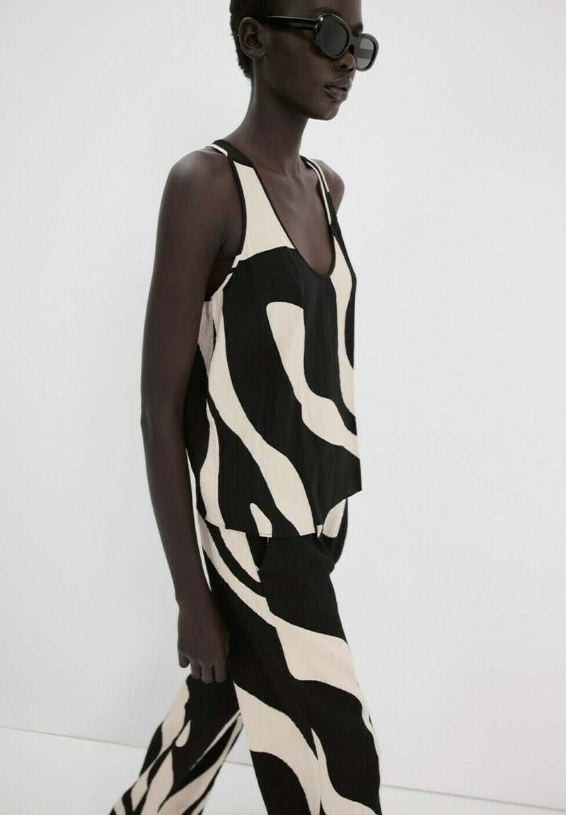 Femeie purtând un top fără mâneci și pantaloni de culoare albă-negru cu model abstract, purtând ochelari de soare mari, negri, mergând pe un fundal simplu.