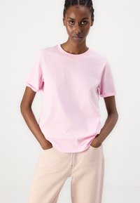 Camiseta de manga corta de color rosa claro, hecha de algodón, con cuello redondo y puños enrollados, combinada con pantalones beige claros.